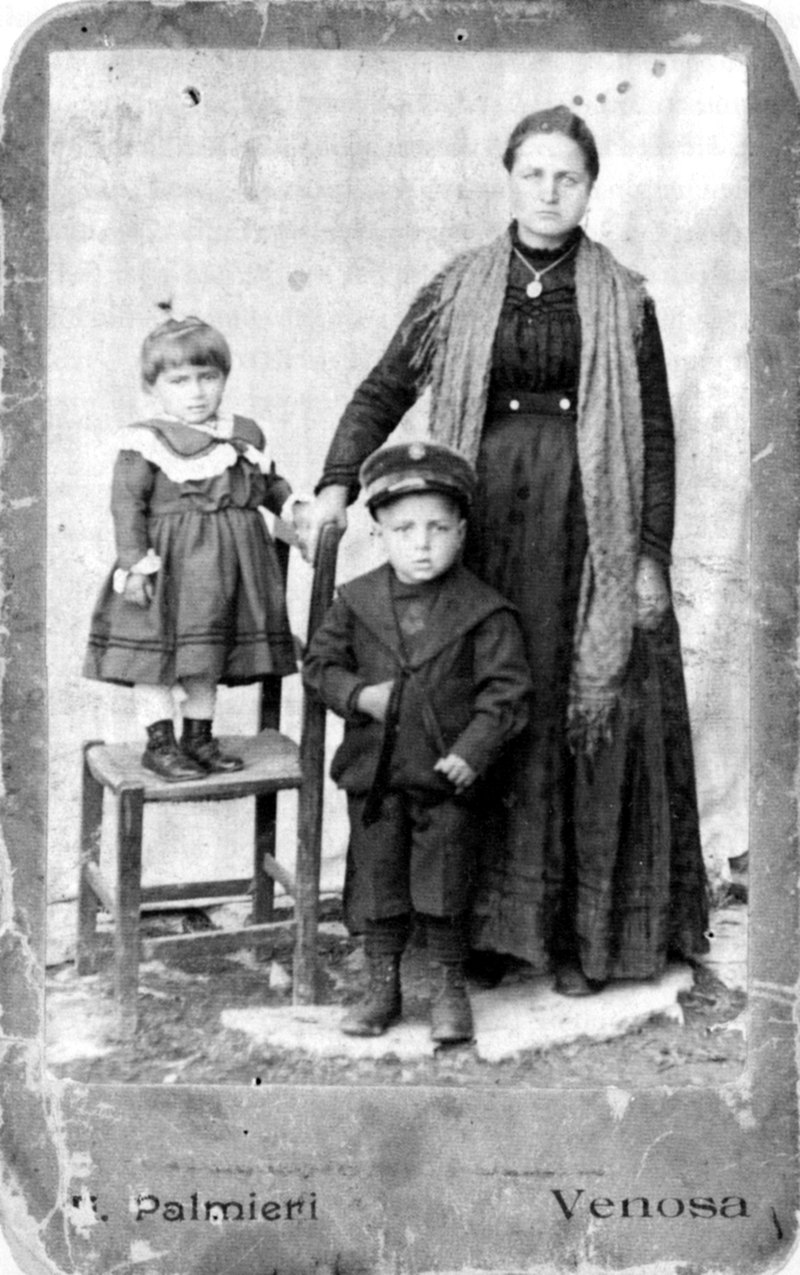 Studio portrait of Maria Joanna La Centra standing beside her son Antonio, with her daughter Elizabetta standing on a chair; the card is stamped Palmieri, Venosa