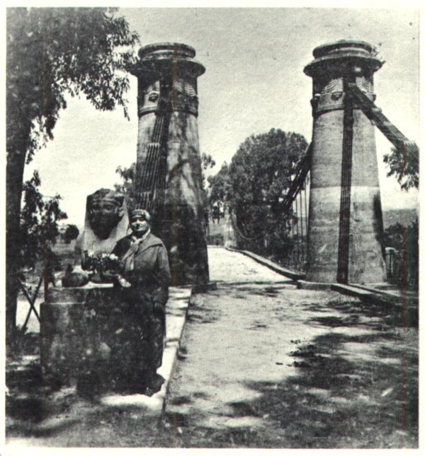 Old black-and-white photograph of the bridge entrance: two tall stone towers with iron chains, a stone sphinx on a pedestal, and a visitor in a long coat standing beside it